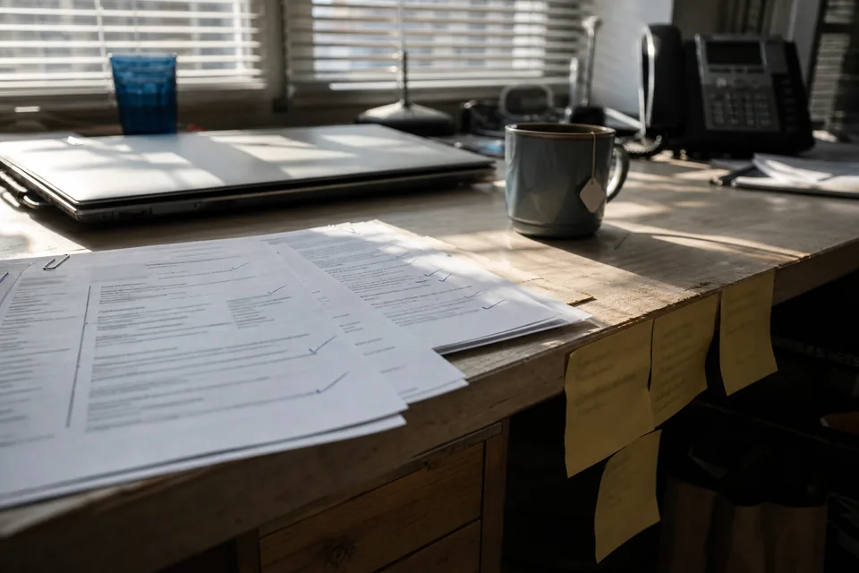 Recruiter desk mid hiring with printed CVs, blue ticks, sticky notes and a half closed laptop