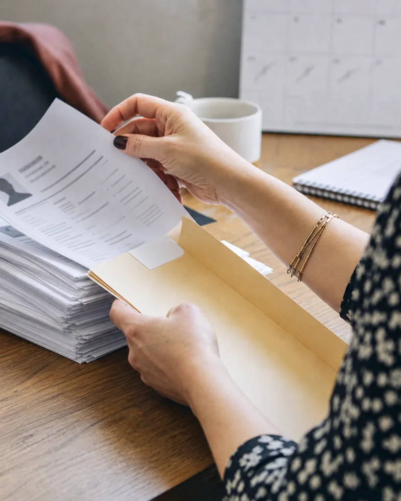 Recruiter moving a few printed candidate profiles from a tall pile into a slim folder, with an interview calendar behind
